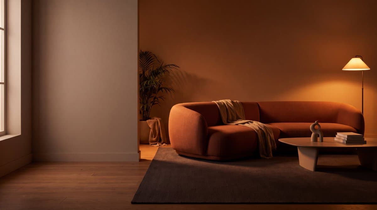 A living room at dusk with a curved rust-coloured sofa, a woven rug and a lit floor lamp